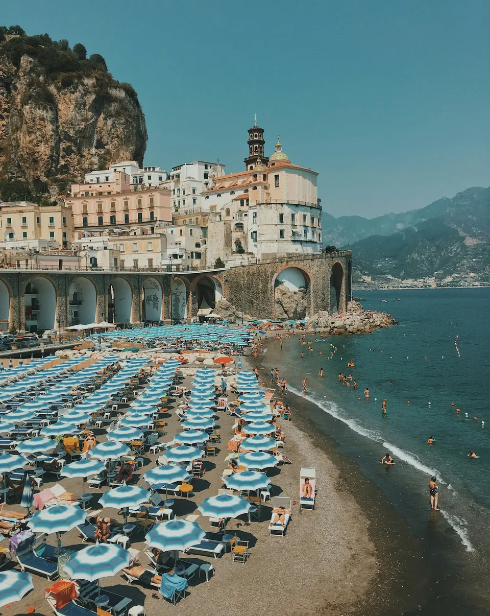 Blick auf die Küste zwischen Amalfi und Atrani mit an den Fels gebauten Häusern, der Straße am Meer und der maiolikierten Kuppel der Kollegiatskirche im Hintergrund